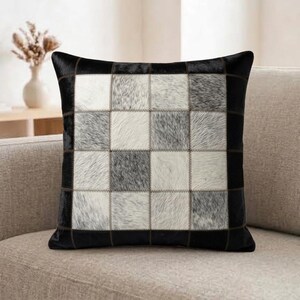 Könnte beinhalten: Ein quadratisches Zierkissen mit einem Patchwork-Design aus Kuhfell-Quadraten in Schwarz-, Weiß- und Grautönen. Die Vorderseite besteht aus einem Gitter aus abwechselnd texturierten Fellstücken, die mit sichtbarem dunklem Garn vernäht sind.