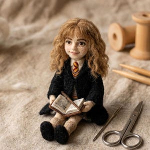 Puede incluir: Una muñeca de lana fieltrada con cabello castaño rizado, que viste una túnica negra sobre un suéter gris y una corbata a rayas rojas y doradas. La muñeca sostiene un pequeño libro abierto y una varita marrón fina, con tez clara y pecas.