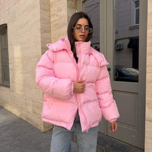 Könnte beinhalten: Eine leuchtend rosa Steppjacke mit hohem Kragen, abnehmbarer Kapuze mit Druckknöpfen, langen Ärmeln mit elastischen Bündchen und einem durchgehenden Frontreißverschluss. Die Jacke wird mit einer hellblauen Jeans und einer silbernen Brille kombiniert.