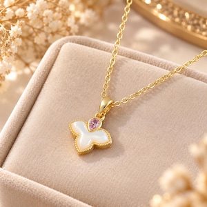 Op de afbeelding: Een goudkleurige ketting met een hanger met een witte parelmoeren inleg in de vorm van een driebladige bloem, geaccentueerd door een kleine, peervormige roze edelsteen aan de bovenkant, hangend aan een fijne goudkleurige schakelketting.