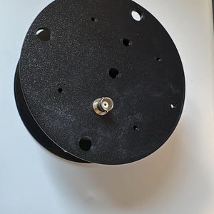 Puede incluir: Una placa de montaje circular negra con acabado texturizado, que presenta varios orificios pretaladrados de diferentes tamaños y un conector BNC de color plateado que sobresale del centro de la superficie plana.