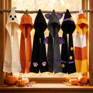 May include: Six crocheted hooded scarves hanging on a wooden branch. The designs include a white ghost, an orange pumpkin, a black scarf with pink paw prints, a dark gray bat with purple wings, a black witch hat with stars, and a candy corn scarf.