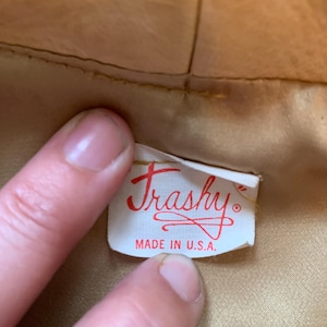 May include: A white rectangular clothing label with red text reading "Trashy" in a cursive script and "MADE IN U.S.A." in block letters, attached to the interior of a tan-colored garment with a smooth, satin-like lining fabric.