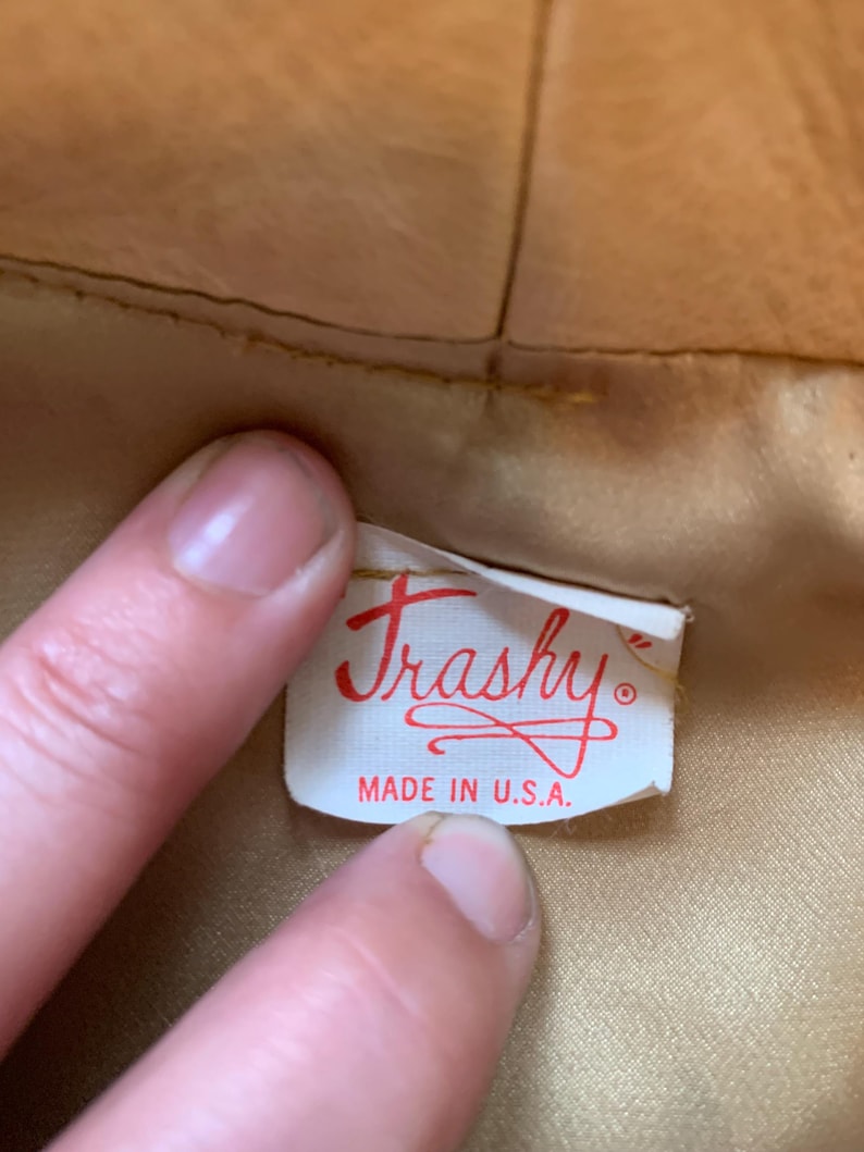 May include: A white rectangular clothing label with red text reading "Trashy" in a cursive script and "MADE IN U.S.A." in block letters, attached to the interior of a tan-colored garment with a smooth, satin-like lining fabric.