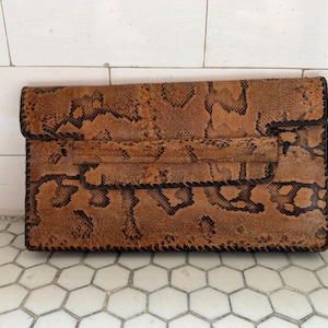 May include: A rectangular brown clutch bag with a snakeskin pattern. The bag has a flap closure and a small handle. The edges are stitched with black thread. The clutch is made of leather and is approximately 30 cm wide and 20 cm tall.