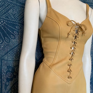 May include: A sleeveless tan leather dress featuring a lace-up front closure with matching leather cord ties. The bodice has a V-neckline with thick shoulder straps and vertical seam detailing that extends down the torso to the waistline.