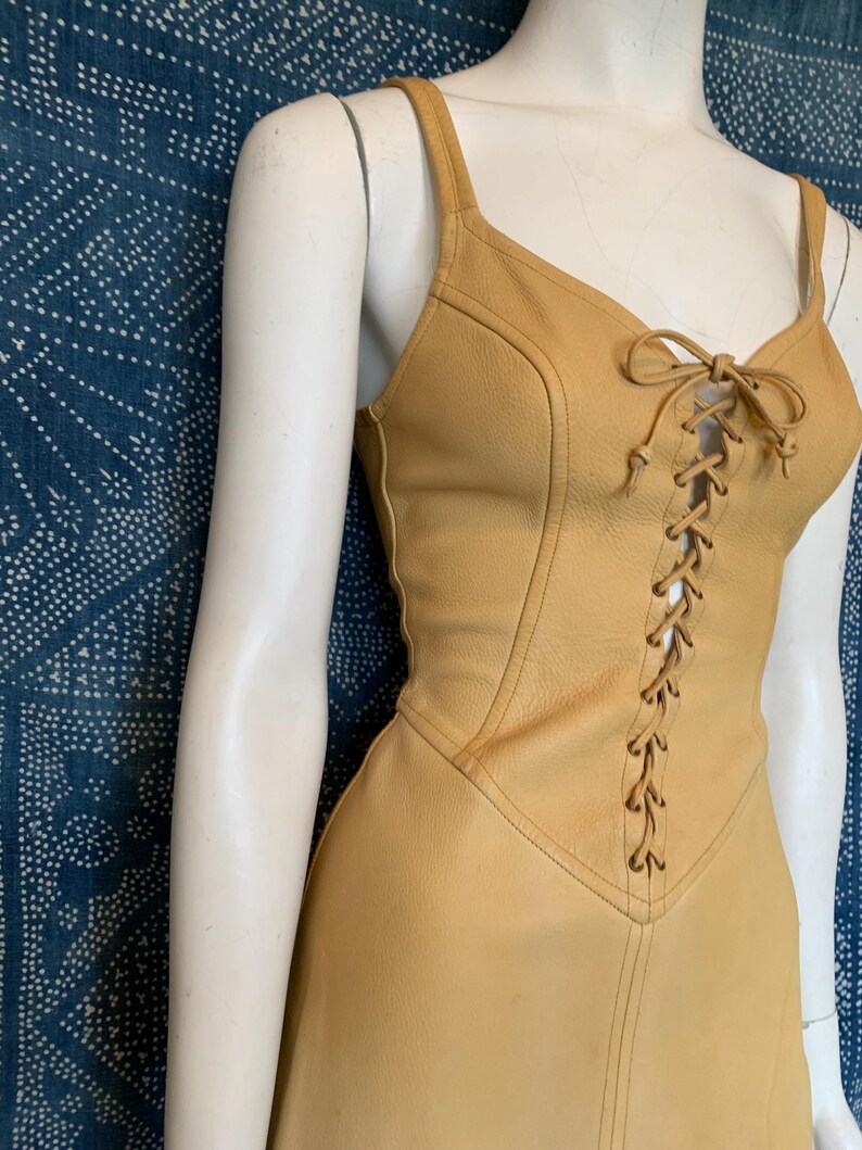 May include: A sleeveless tan leather dress featuring a lace-up front closure with matching leather cord ties. The bodice has a V-neckline with thick shoulder straps and vertical seam detailing that extends down the torso to the waistline.