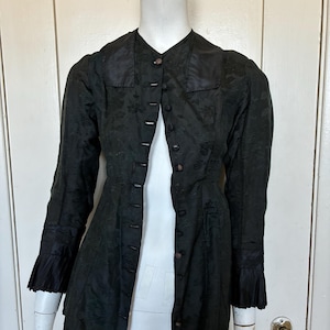 Peut inclure: Une veste noire à manches longues avec un motif floral jacquard ton sur ton, un col haut et une fermeture boutonnée sur le devant avec de petits boutons ronds. Les manches sont terminées par des poignets plissés et la coupe est ajustée.