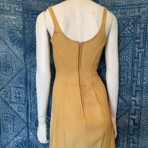 May include: A mustard yellow sleeveless dress featuring a scoop neckline, a fitted bodice with vertical seam detailing, and a central vertical zipper closure running down the back. The garment is displayed on a mannequin, showcasing a structured silhouette.