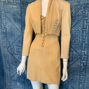 May include: A tan leather two-piece set consisting of a cropped jacket and a sleeveless mini dress. The jacket features long sleeves and a lace-up detail on the front. The dress has a matching lace-up bodice with a central vertical seam.