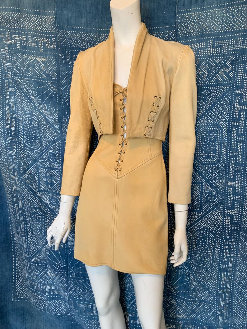 May include: A tan leather two-piece set consisting of a cropped jacket and a sleeveless mini dress. The jacket features long sleeves and a lace-up detail on the front. The dress has a matching lace-up bodice with a central vertical seam.