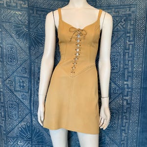 May include: A tan sleeveless mini dress featuring a lace-up front closure with matching tan cord ties. The dress has a fitted bodice with vertical seam detailing and a slightly flared skirt, displayed on a mannequin against a blue patterned backdrop.