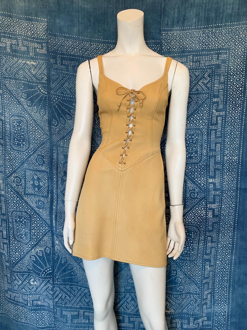 May include: A tan sleeveless mini dress featuring a lace-up front closure with matching tan cord ties. The dress has a fitted bodice with vertical seam detailing and a slightly flared skirt, displayed on a mannequin against a blue patterned backdrop.
