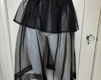 May include: A black sheer mesh skirt featuring a layered design with a shorter, pleated upper section and a longer, flowing lower panel. The garment has a solid black waistband and a reinforced hemline at the bottom of the sheer fabric.