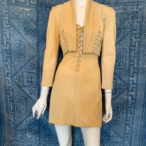 May include: A light tan leather two-piece set consisting of a cropped jacket with long sleeves and a matching mini dress. Both pieces feature vertical lace-up detailing with metal grommets running down the center front of the bodice and jacket.