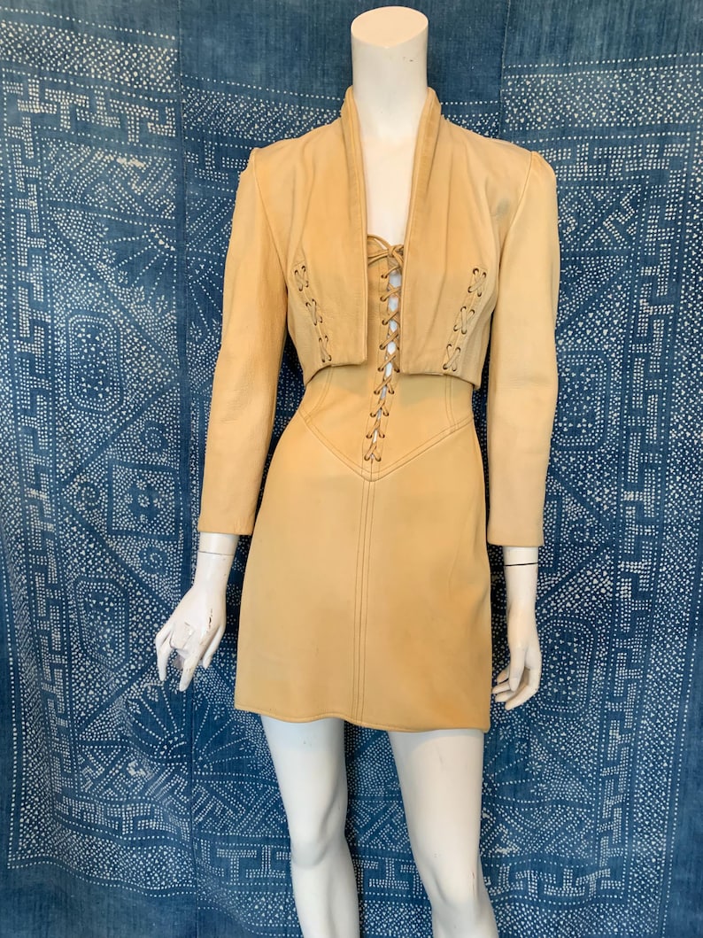 May include: A light tan leather two-piece set consisting of a cropped jacket with long sleeves and a matching mini dress. Both pieces feature vertical lace-up detailing with metal grommets running down the center front of the bodice and jacket.