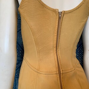 May include: A sleeveless mustard yellow leather bodice featuring a textured surface, a scoop neckline, and a vertical center back zipper closure. The garment is constructed with multiple contoured panels and visible stitching throughout the design.