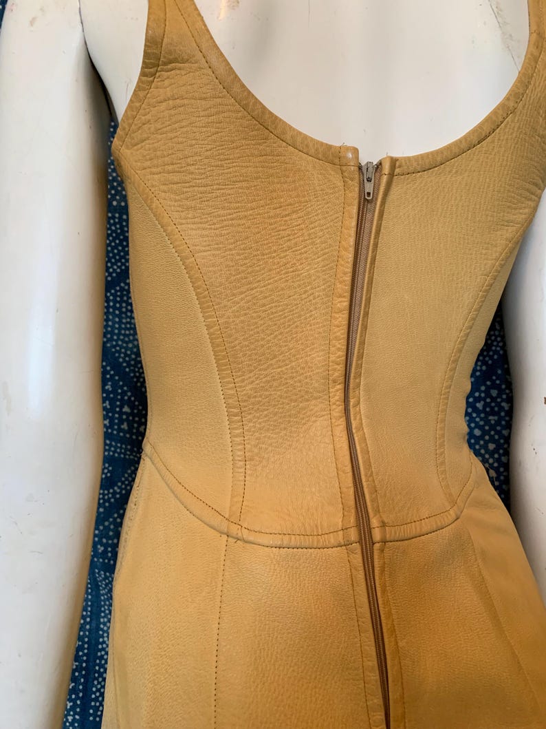 May include: A sleeveless mustard yellow leather bodice featuring a textured surface, a scoop neckline, and a vertical center back zipper closure. The garment is constructed with multiple contoured panels and visible stitching throughout the design.