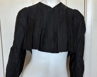 May include: A black cropped jacket featuring a round neckline, long sleeves, and vertical pleats across the front bodice. The garment has a structured shoulder design and a straight hemline that ends above the waist, displaying a solid dark color.