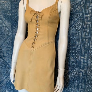 May include: A mustard yellow sleeveless mini dress featuring a lace-up front closure with matching cord ties, a V-neckline, and vertical seam detailing that runs from the bodice down to the hemline, creating a structured and fitted silhouette.