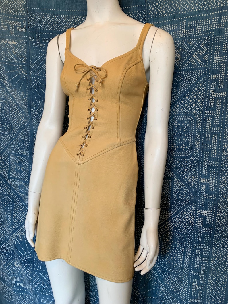 May include: A mustard yellow sleeveless mini dress featuring a lace-up front closure with matching cord ties, a V-neckline, and vertical seam detailing that runs from the bodice down to the hemline, creating a structured and fitted silhouette.