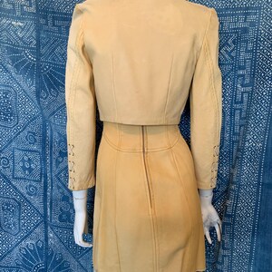 May include: A light tan leather two-piece set consisting of a cropped jacket and a matching skirt. The jacket features long sleeves with decorative lace-up detailing on the forearms. The skirt has a vertical center back zipper closure.