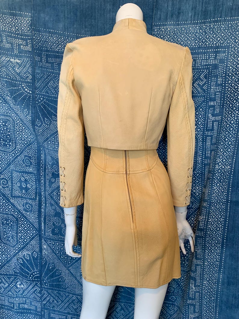 May include: A light tan leather two-piece set consisting of a cropped jacket and a matching skirt. The jacket features long sleeves with decorative lace-up detailing on the forearms. The skirt has a vertical center back zipper closure.