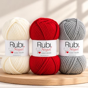 Puede incluir: Tres ovillos de lana Rubi Nogal en color crema, rojo y gris. Cada ovillo tiene una etiqueta de papel blanco con el texto Rubi Nogal, un icono de oveja, un corazón rojo, el texto nueva edición y la web lanasrubi.com.