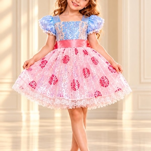 Puede incluir: Un vestido con corpiño azul claro con superposiciones de encaje, mangas abullonadas y una cintura de satén rosa. La falda es rosa con lunares de lentejuelas y un dobladillo de encaje blanco. Incluye una diadema a juego con orejas negras y lazo.