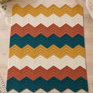 May include: A rectangular crochet blanket featuring a repeating chevron pattern in horizontal stripes of mustard yellow, cream, terracotta orange, and deep teal. The textile has a textured, stitched surface with a zigzag edge along the top and bottom.