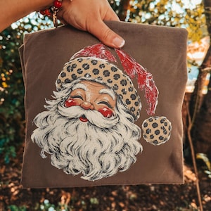 May include: A brown t-shirt featuring a graphic print of a Santa Claus face with a white beard, rosy cheeks, and a hat decorated with a tan and black leopard print pattern and a red textured accent, including a matching leopard print pom-pom.