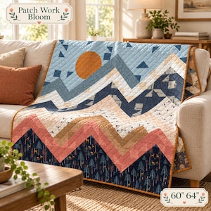 May include: A quilt featuring a mountain landscape with a large orange sun, blue sky, and dark blue forest base with deer silhouettes. The design uses geometric chevron patterns in shades of blue, pink, and tan. The quilt measures 60 by 64 inches.
