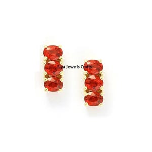 May include: A pair of gold stud earrings, each featuring three vertically aligned oval-shaped red gemstones held by small prongs. The text Siya Jewels Crafts is visible between the two earrings against a plain white background.