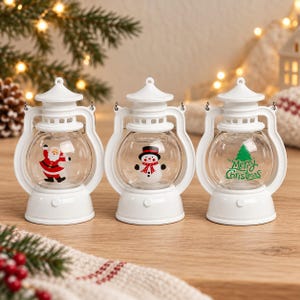 Peut inclure: Trois lanternes en plastique blanc avec des centres en verre transparent. La lanterne de gauche présente un motif de Père Noël, celle du centre un bonhomme de neige avec une écharpe rouge, et celle de droite un sapin vert avec Merry Christmas.
