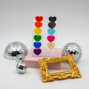 May include: Two pairs of heart-shaped earrings in a rainbow of colours, displayed on a clear stand. The earrings are red, orange, yellow, green, blue, and purple. The other pair is black, brown, light blue, pink, and cream. Disco balls and a gold frame are in the background.