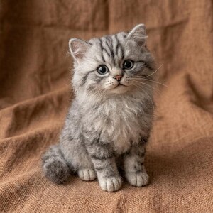 Puede incluir: Una figura de gatito de lana fieltrada con un pelaje esponjoso gris y blanco con rayas atigradas oscuras. La pequeña escultura tiene grandes ojos oscuros, una nariz rosada y largos bigotes blancos, capturando una pose sentada.