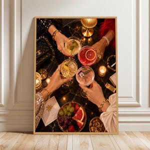 Op de afbeelding: Vier handen houden verschillende cocktails in glazen vast, waaronder drankjes met een schijfje grapefruit, een olijf en een bloemgarnering. De scène toont gouden armbanden, een zwarte gewatteerde handtas, een schaal met druiven en bessen, en kleine brandende kaarsjes op een zwarte marmeren tafel.