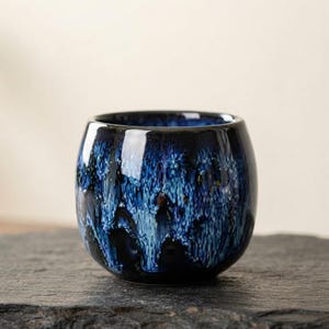 May include: A rounded ceramic cup featuring a deep blue and black glaze with a mottled, drip-like pattern. The exterior surface displays a glossy finish with varying shades of cobalt and navy blue, creating a textured visual effect across the body.