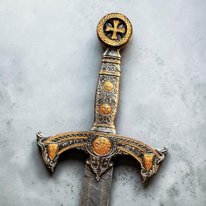 May include: A decorative sword featuring an ornate silver and gold hilt with intricate engravings. The pommel displays a cross emblem, while the crossguard is adorned with circular gold medallions and shield-shaped accents.