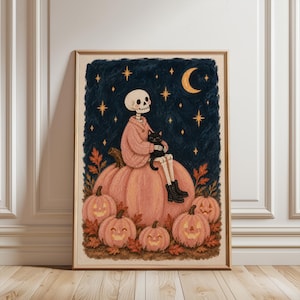 Op de afbeelding: Een ingelijste kunstdruk met een skelet in een roze trui en zwarte laarzen, zittend op een grote roze pompoen terwijl het een zwarte kat vasthoudt. De scène bevat kleinere pompoenlantaarns, herfstbladeren en een maansikkel in een donkere lucht.