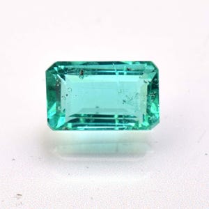 May include: A rectangular emerald-cut gemstone with a vibrant teal-green color. The stone features faceted surfaces that reflect light, showing internal clarity characteristics and a polished finish with sharp, defined edges.
