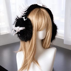 May include: Black faux fur earmuffs featuring a white, skeletal-inspired spine and rib-like structure along the top and sides. The white, bony-textured accents contrast against the soft, dark, plush material of the ear coverings.