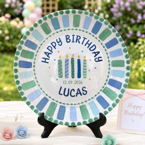 May include: A round ceramic plate featuring a border of alternating blue and green rectangular brushstrokes. The center displays the text Happy Birthday, five colorful candles, the date 12.09.2026, and the name LUCAS in blue lettering.