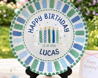May include: A round ceramic plate featuring a border of alternating blue and green rectangular brushstrokes. The center displays the text Happy Birthday, five colorful candles, the date 12.09.2026, and the name LUCAS in blue lettering.