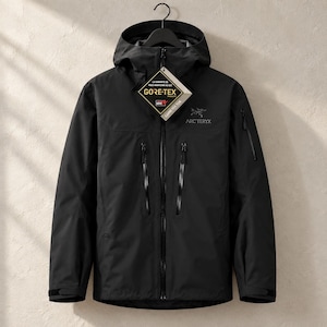 Op de afbeelding: Een zwarte jas met capuchon, voorzien van een volledige ritssluiting aan de voorzijde, twee verticale borstzakken met gesealde ritsen en een mouwzak. Op de borst staan het Arc'teryx-logo en een Gore-Tex productlabel.