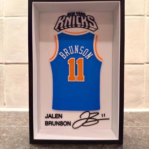 May include: A framed 3D wall art piece featuring a blue New York Knicks jersey with the name BRUNSON and number 11 in orange and white. Below the jersey, the text JALEN BRUNSON is printed next to a black signature and the number 11.