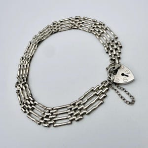 Puede incluir: Una pulsera de tono plateado con un diseño de cadena de múltiples hilos y un dije de candado en forma de corazón. El candado incluye una cerradura central y detalles de tornillos decorativos, con una pequeña cadena de seguridad adjunta.