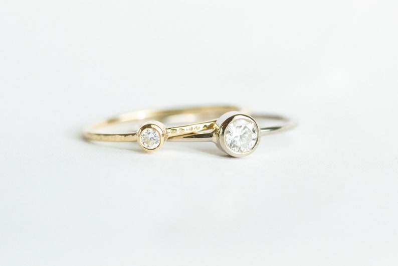 May include: Two delicate rings with clear gemstones. One ring is gold with a smaller stone, and the other is silver with a larger, bezel-set stone. The rings are set against a white background, highlighting their simple elegance.