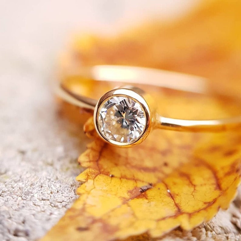 May include: A gold ring with a round, clear gemstone set in a bezel setting. The ring is resting on a yellow leaf, with a blurred background. The ring's band is thin and delicate, and the gemstone sparkles in the light.