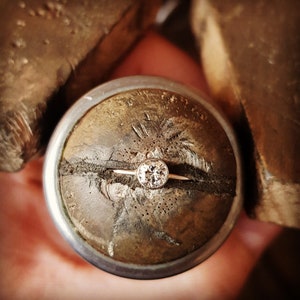 May include: A gold ring with a large, round, clear diamond set in a bezel setting. The ring is on a brown, textured surface, likely a jeweler's tool. The background is blurred, suggesting a close-up shot of the jewelry.